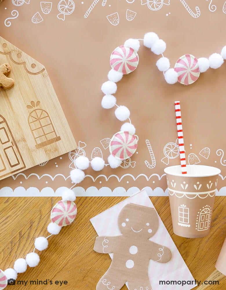 A tabletop featuring Momo Party's gingerbread man table runner, on top of it are the 4.25" x 7.75" gingerbread man shaped napkins by My Mind's Eye, along with a bamboo gingerbread house shaped serving tray which has two cute gingerbread man cookies on tis, next to it lay a pink candy cane felt banner which is surrounded with two gingerbread house party cups and pink striped dinner napkins from Momo Party for a kid's cookie decorating / gingerbread house building party.