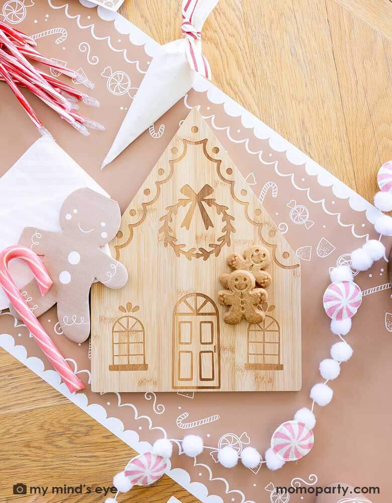 A tabletop featuring Momo Party's gingerbread man table runner, on top of it are the 4.25" x 7.75" gingerbread man shaped napkins by My Mind's Eye, along with a bamboo gingerbread house shaped serving tray which has two cute gingerbread man cookies on tis, next to it lay a pink candy cane felt banner which is surrounded with candy canes and cookie decoration butter cream piping bags for a kid's cookie decorating / gingerbread house building party.