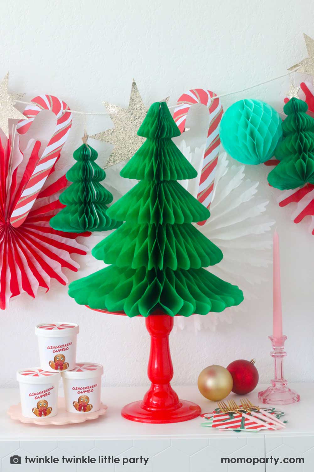 A Christmas Dessert Table decorated with Momo Party's Christmas honeycomb decorations featuring candy canes, honeycomb Christmas trees and mint honeycomb balls. On the table there's a red cake stand with Momo Party's 17" light green honeycomb Christmas tree on it, a pink candle on a clear pink candle stand next to Meri Meri's present with a bow shaped napkins in festive colors of red and green. On the left, there are some peppermint treat boxes stacked, making this a perfect Holiday party inspo.