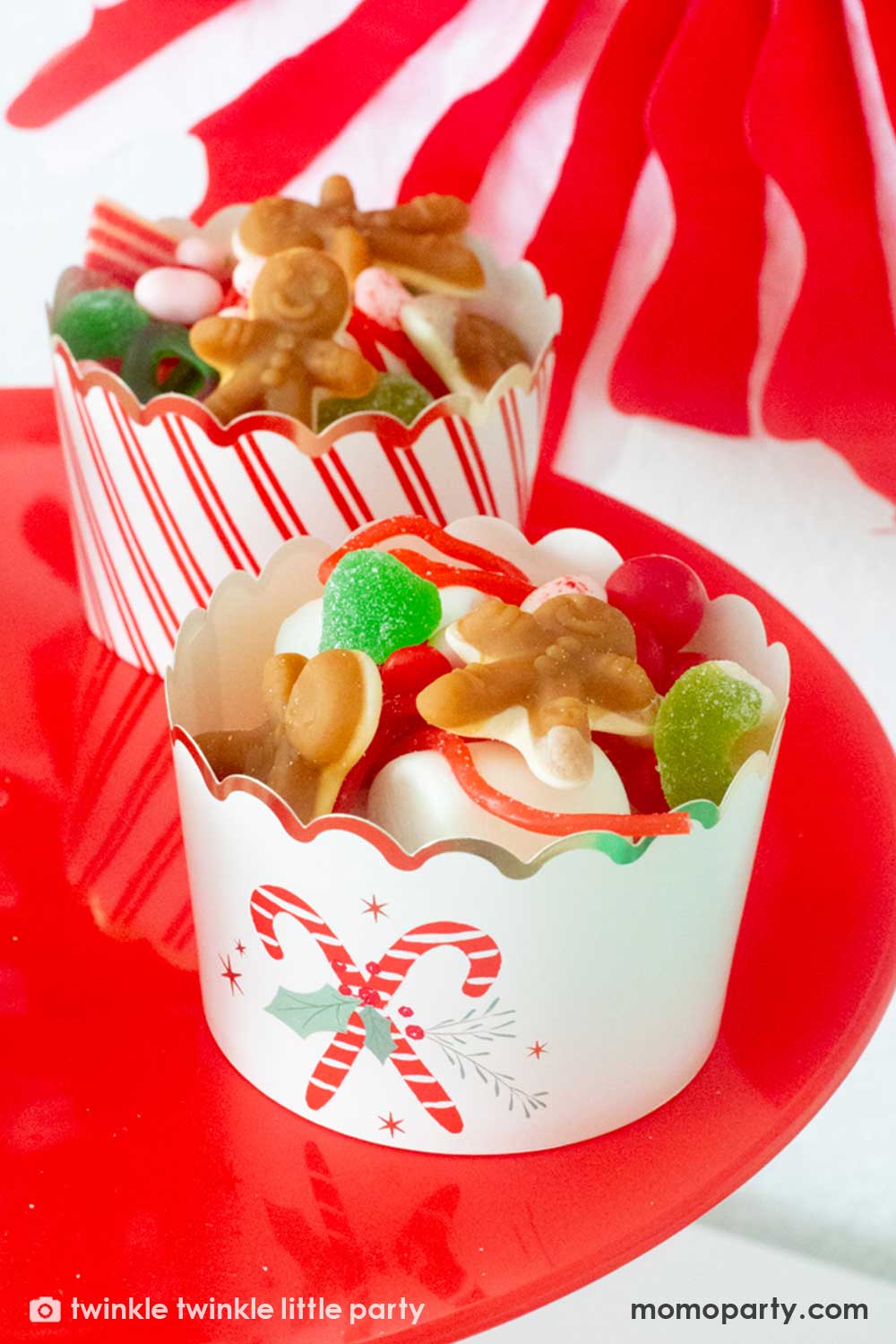 A close up shot of Christmas Dessert Table decorated with Momo Party's Christmas honeycomb decorations featuring candy canes, honeycomb Christmas trees and mint honeycomb balls. On the table there's a red cake stand with Momo Party's Candy Cane Jumbo food cups filled with lots of Holiday teats including green candies, gingerbread man gummies and red gum balls, making this a great idea for festive Holiday party treat and snack ideas for kids.
