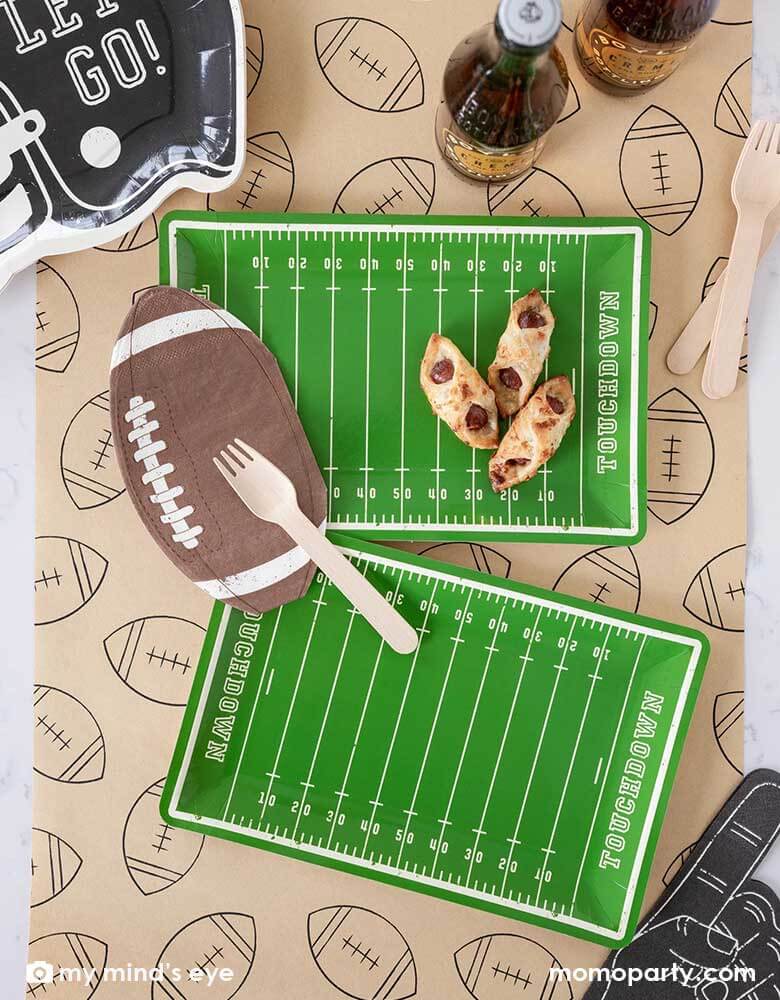 A football party table features Momo Party's Football Field shaped plates, on the plate there are some party snacks and around the plates there are Momo Party's football shaped napkins, black helmet shaped plates featuring "Let's Go"" on it, and a no #1 finger shaped black napkin on a light brown table runner with football illustration on it. With party drinks around, it makes a great party table decoration for a fun football season watch game party or a Super Bowl party!