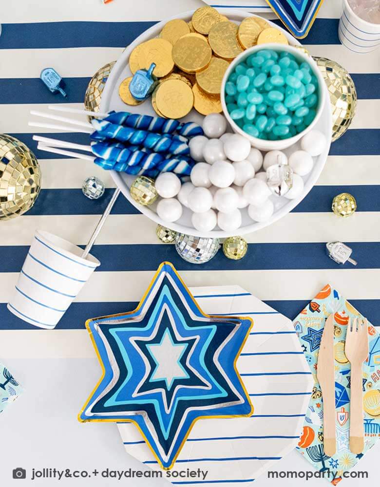 A festive Hanukkah party table featuring Momo Party's blue striped plates and party cups with Festival of Lights small star shaped plates and patterned napkins by Daydream Society. On the a blue striped table runner is a cupcake holder with treats and candies in coordinating blue and white colors and some coin chocolates and Dreidel candies, makes it a great inspo for a festive Hanukkah party for family gathering. 