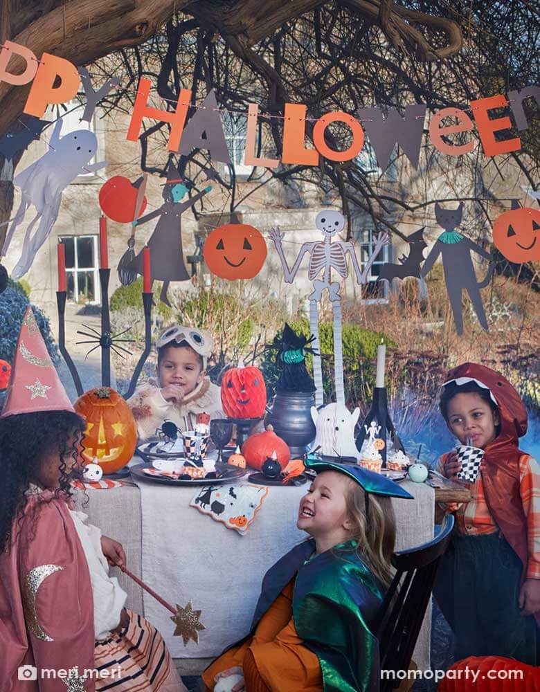 A fun Halloween scene with a party table filled with festive Halloween themed party supplies by Momo Party including Halloween cupcake toppers, Halloween plates, cups, napkins and Halloween character honeycomb by Meri Meri. On the top, there's a 7 ft "Happy Halloween"" party garland hung featuring iconic Halloween characters including skeletons, witches, ghosts, black cats and pumpkins. A few kids dressed up in costumes sitting and having fun around the party table.