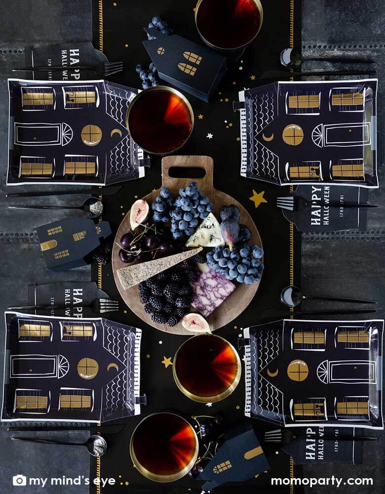 A black and gold Halloween party table featuring Momo Party's 8" x 10.5" haunted village haunted house shape plates and black tombstone shaped napkins with Happy Halloween words on them. Around the tableware, there are red drinks and black themed charcuterie board on Momo Party's mystical dark night table runner, gives a spooky vibe to an elegant Halloween tablescape.