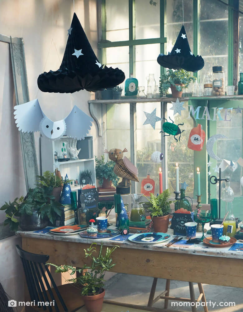 A witch and wizard party table set up featuring Momo Party's making magic party supplies collection by Meri Meri including hanging witch hat honeycomb decorations, potion bottle shaped plates, wizard spell book napkins, witch cauldron shaped party bags, owl honeycomb decoration, night ombre napkins and cups with moon and stars on them, and bug confetti scattered on the table, makes it a great inspiration for kid's harry potter themed party or a Halloween witch and wizard themed bash.