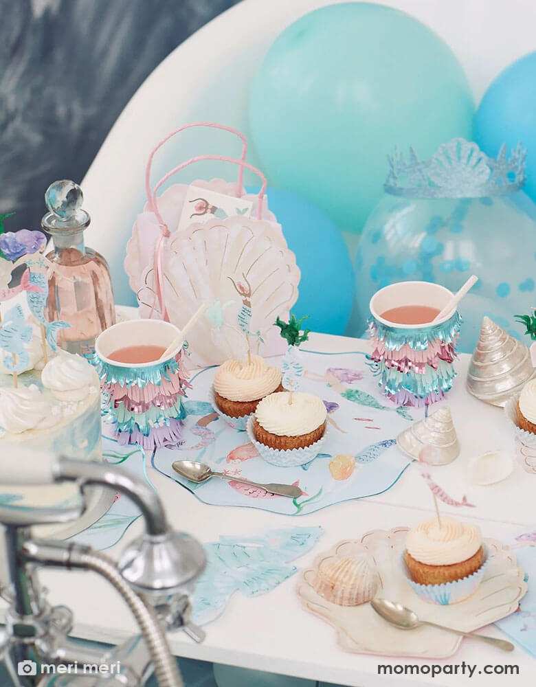 A beautiful kid's under the sea mermaid themed party table featuring Momo Party's mermaid party supplies including plates, iridescent fringe party cups, shell shaped plates, seahorse shaped napkins and pink shell shaped party bags by Meri Meri. In the back there are some fun latex balloons in light blue shades. On the table there are some cupcakes topped with mermaid themed cupcake toppers, makes this a great inspo for a whimsy mermaid party for girls.