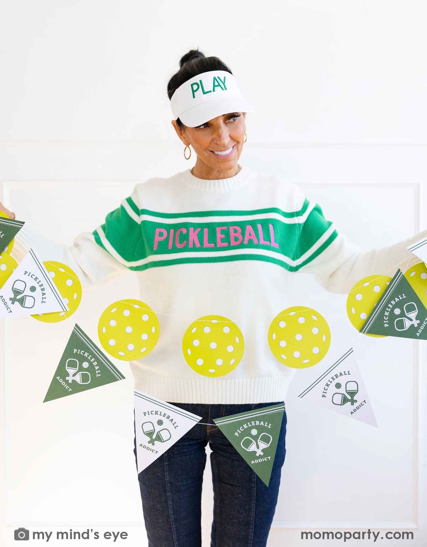 A lady wearing a 'Pickleball' themed sweater and pants with her white hat says "Play" holding Momo Party's pickleball party banner set by My Mind's Eye on a white background.