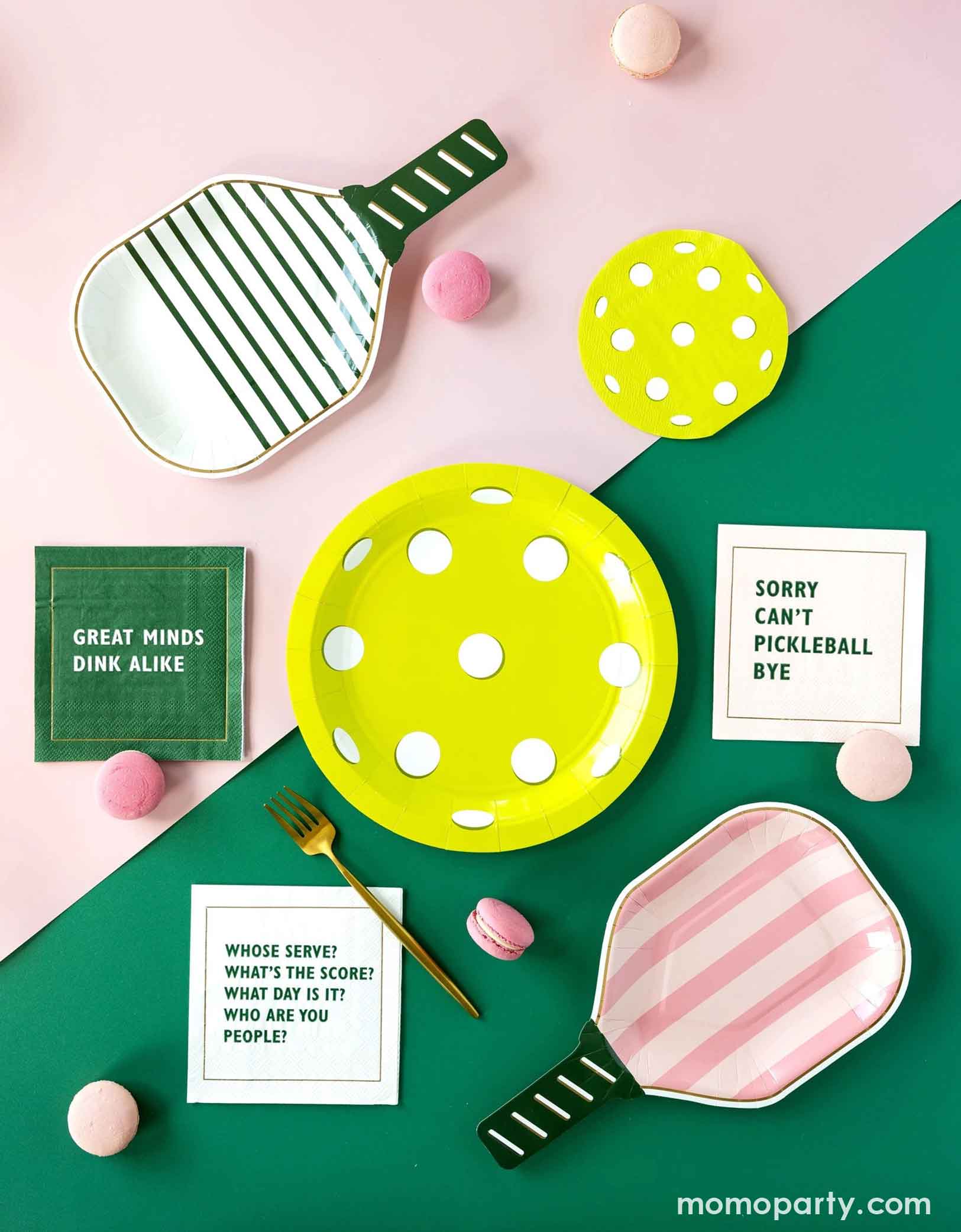An overhead shot of pink and green table with pickleball-themed party supplies from Momo Party including pickleball paddle shaped plates, pickleball shaped plate and matching napkin, and pickleball sayings napkins in pink, white and green by My Mind's Eye. Along with some pink macarons and a golden fork - making this an inspiration for a pickleball themed celebration.