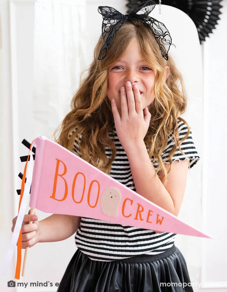 A girl in her Halloween black outfit holding Momo Party's 14" x 15" pink Boo Crew felt party pennant by My Mind's Eye. In a pink Halloween hue, this felt party pennant with an adorable ghost illustration on it and orange and black ribbons, it will make sure your ghouls get the recognition they deserve. This Boo Crew felt pennant is perfect to set a scene for your girl's pink Halloween bash.