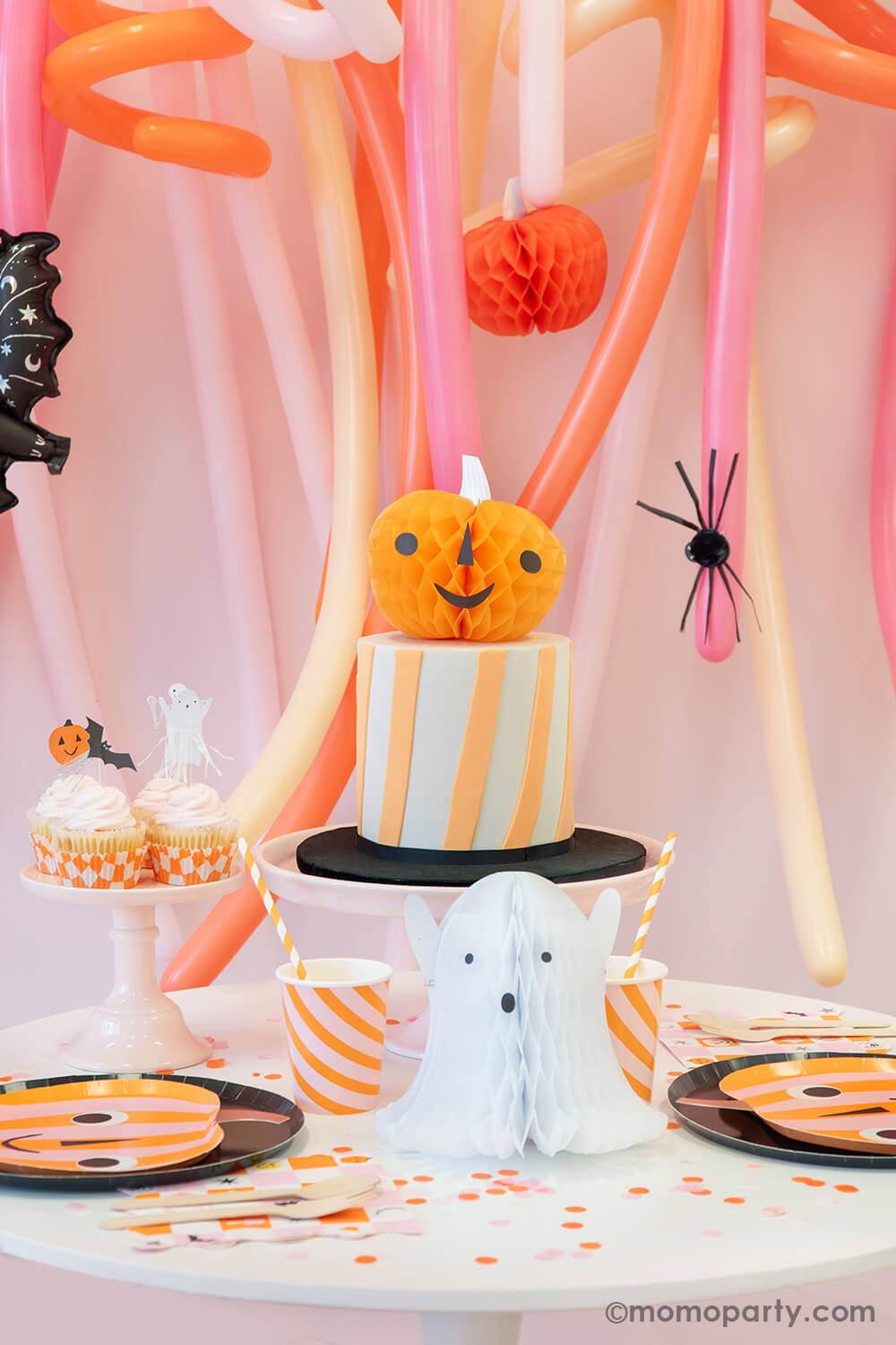 Momo Party's Trick or Treat Pink Halloween box set up featuring a festive long balloon backdrop with curling long balloons in coral, pink, rose and peach colors. On the balloon backdrop it's decorated with paper pumpkin honeycombs and a mini bat foil balloon. In front of the balloons, a small round table is filled with Halloween party supplies including pumpkin shaped plates, a bat garland, ghost and pumpkin honeycomb decorations from momoparty.com.