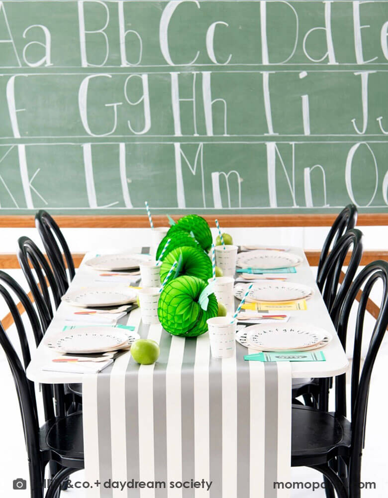 A back to school party table set up in front of a large school chalkboard featuring Momo Party's School Days large and small Plates, along with crayon shaped napkins by Daydream Society. In the middle of the table were some lime green apple honeycombs and green balloons as the centerpiece. All in all makes a great inspiration of a school themed party or first day of school celebration!