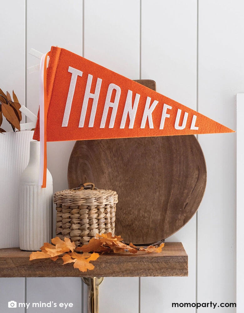 A fall/autumn wooden shelf decorated with a wooden cutting board, a weaved jar, some fall leaves and a couple white vase which one is adorned with Momo Party's 14" orange Thankful felt party pennant by My Mind's Eye.