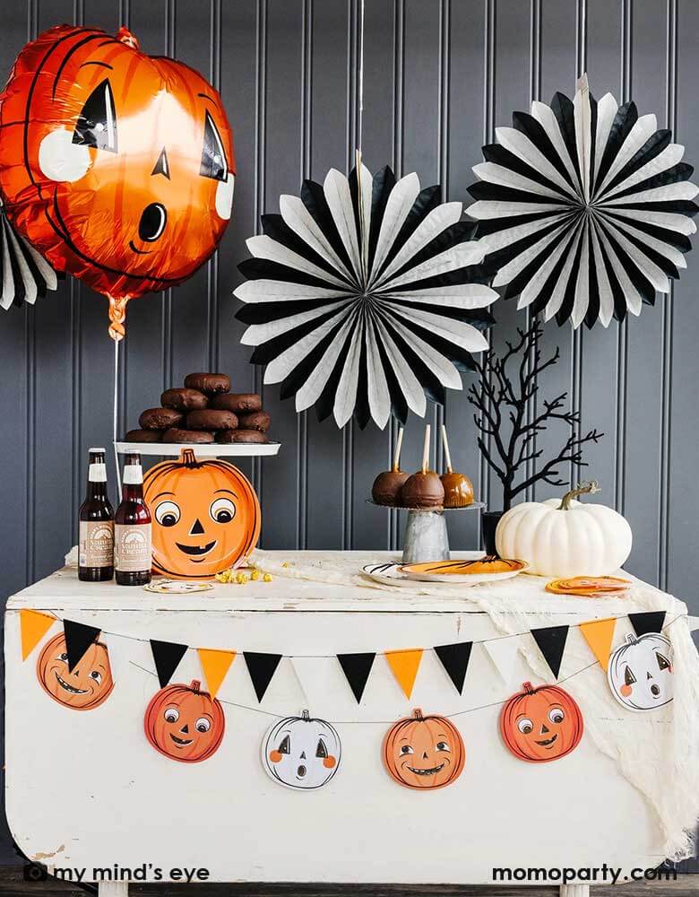 A vintage Halloween table decorated with vintage Halloween styled pumpkin shaped plates, root beers, a white pumpkin, a creepy tree, and some caramel apples. In the back the wall is decorated with My Mind's Eye's oversized black and white tissue paper fans and a vintage styled Halloween foil balloon . All makes a simply yet iconic Halloween decorating inspo this Halloween season.