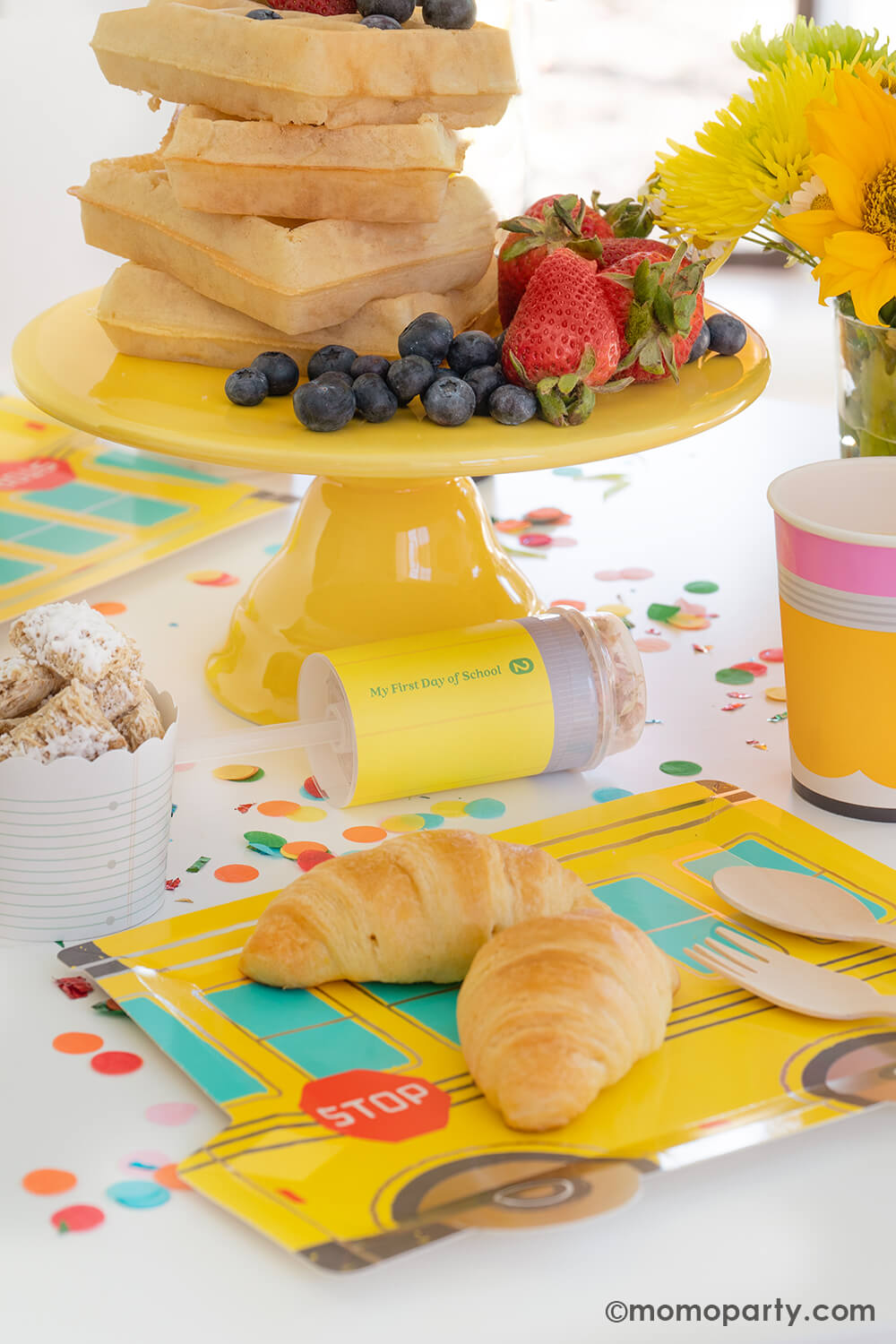 A first day of school breakfast celebration table with delicious breakfast food including Belgium waffles stacked as a cake with mixed berries, on the table is a school bus shaped party plates with mini croissants for kids, along with pencil shaped party cups and pencil party poppers, these back to school party ideas by Momo Party make a great inspiration for any school themed celebrations with your kids at home.