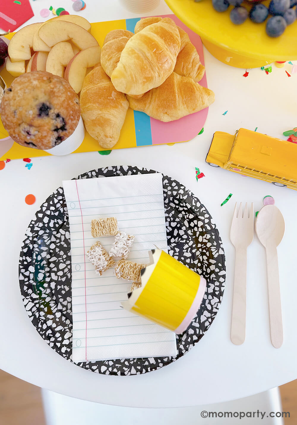 A kid's first day of school celebration party table by Momo Party featuring school themed tableware including art book inspired round plates, notebook napkins and pencil shaped food cups. In the middle, a pencil shaped charcuterie board filled with delicious breakfast items including mini croissants, apple slices, muffles and fruits - a great and healthy back to school breakfast items for moms and can be easily recreated at home.