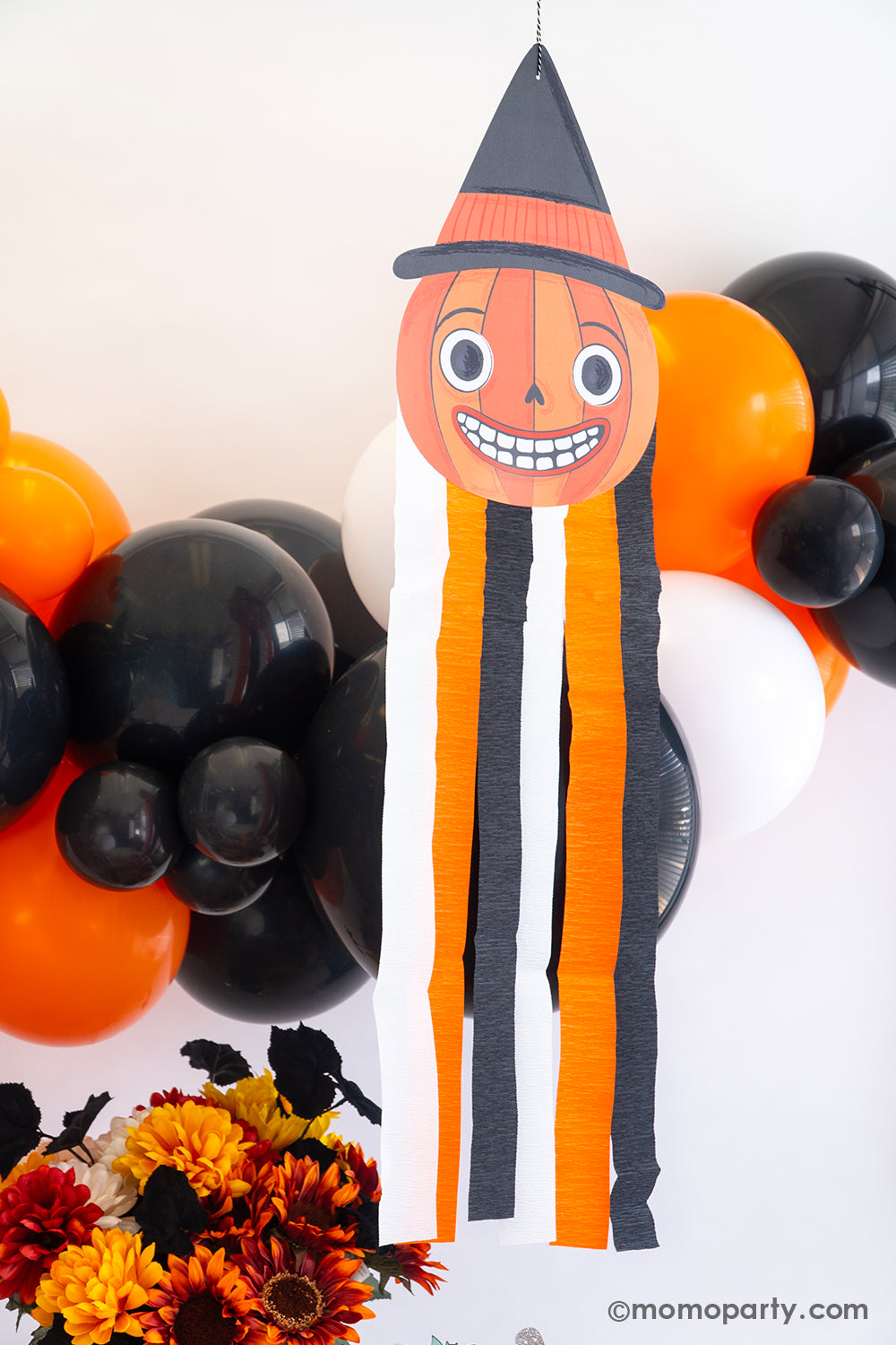 A close-up shot of a Vintage Halloween decorations featuring Momo Party's Halloween character pumpkin hanging decoration with streamers along with a balloon garland in classic Halloween colors of black, orange and white. On the day is a fall flower arrangement with matching colors, making this a great inspo for a simple and modern Halloween bash decorating idea that's also kid's friendly.