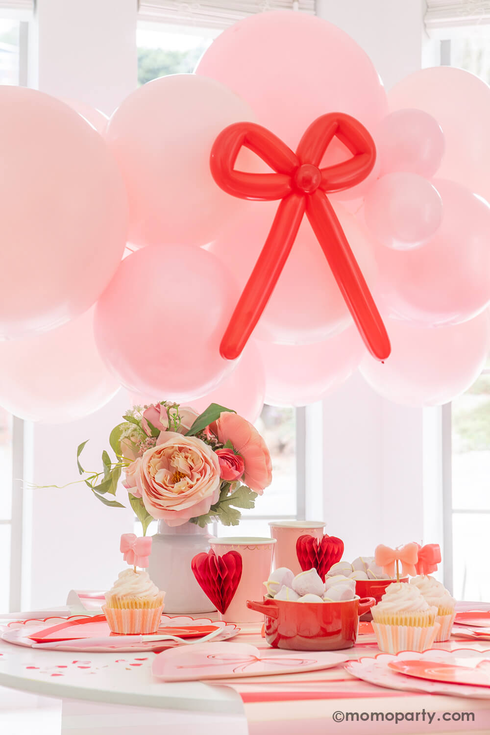A close up shot of Momo Party's Bow-tiful Valentine's Day Party Box for 8 guests. Featuring a pink balloon garland with red bow balloons on it, heart shaped tableware including plates, napkins and party cups, and a beautiful flower bouquet, this adorable party box in classic Valentine's colors of red and pink is perfect for your Valentine's Day party or Galentine's Day celebration.