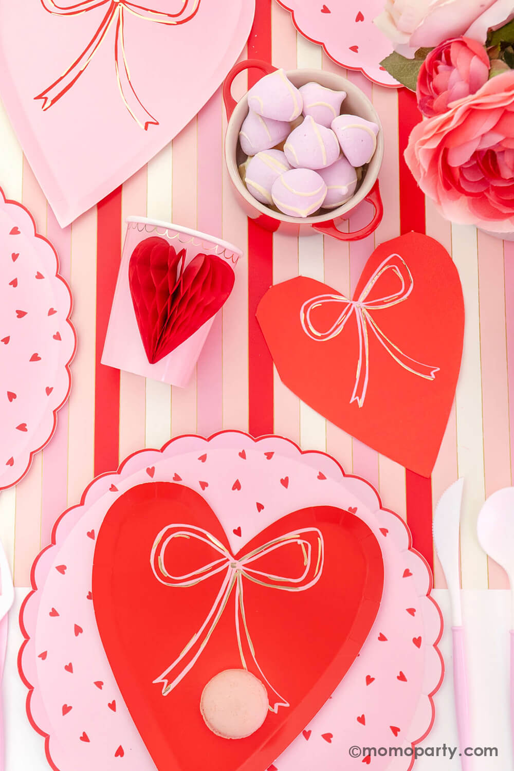 A close-up top view of Momo Party's sweet Pink and Red Bow-Themed Valentine's Day Party table. Featuring a delectable macaron on the Meri Meri Heart With Bow Plates layered with Heart Pattern Dinner Plates, Heart With Bow Napkins, Honeycomb Heart Cups, Blush and White Cutlery Set, Pink Bow Candles atop cupcakes, and sweets in the red Mini Cocotte. All elegantly arranged on a red-striped paper runner. Such a cute idea for elevating your Valentine's celebration or Galentine's Day with your besties.