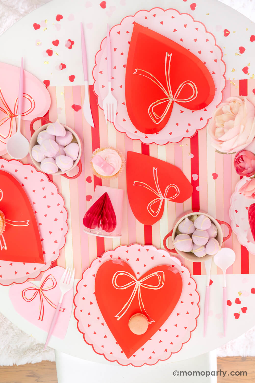 Top view of Momo Party's sweet Pink and Red Bow-Themed Valentine's Day Party table. Featuring Meri Meri Heart With Bow Plates layered with Heart Pattern Dinner Plates, Heart With Bow Napkins, Honeycomb Heart Cups, Blush and White Cutlery Set, Pink Bow Candles atop cupcakes, and sweets in the red Mini Cocotte. All elegantly arranged on a red-striped paper runner, sprinkled around Kisses Artisan Confetti. Such a cute idea for elevating your Valentine's celebration or Galentine's Day with your besties