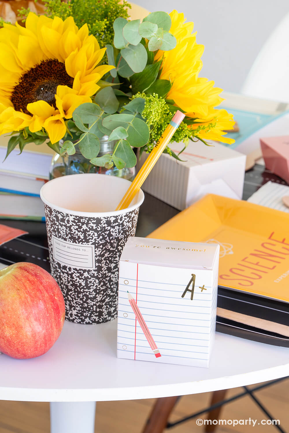An close-up shot of a back to school party table featuring multiple school themed party goods from Momo Party, including text book shaped plates in different colors, composition party cups with pencil straws, and ruled notebook treat boxes, all on the chalkboard table runner. With apples and a sunflower floral arrangement on the table, this makes a perfect idea for your child's back to school party or first day of school celebration!