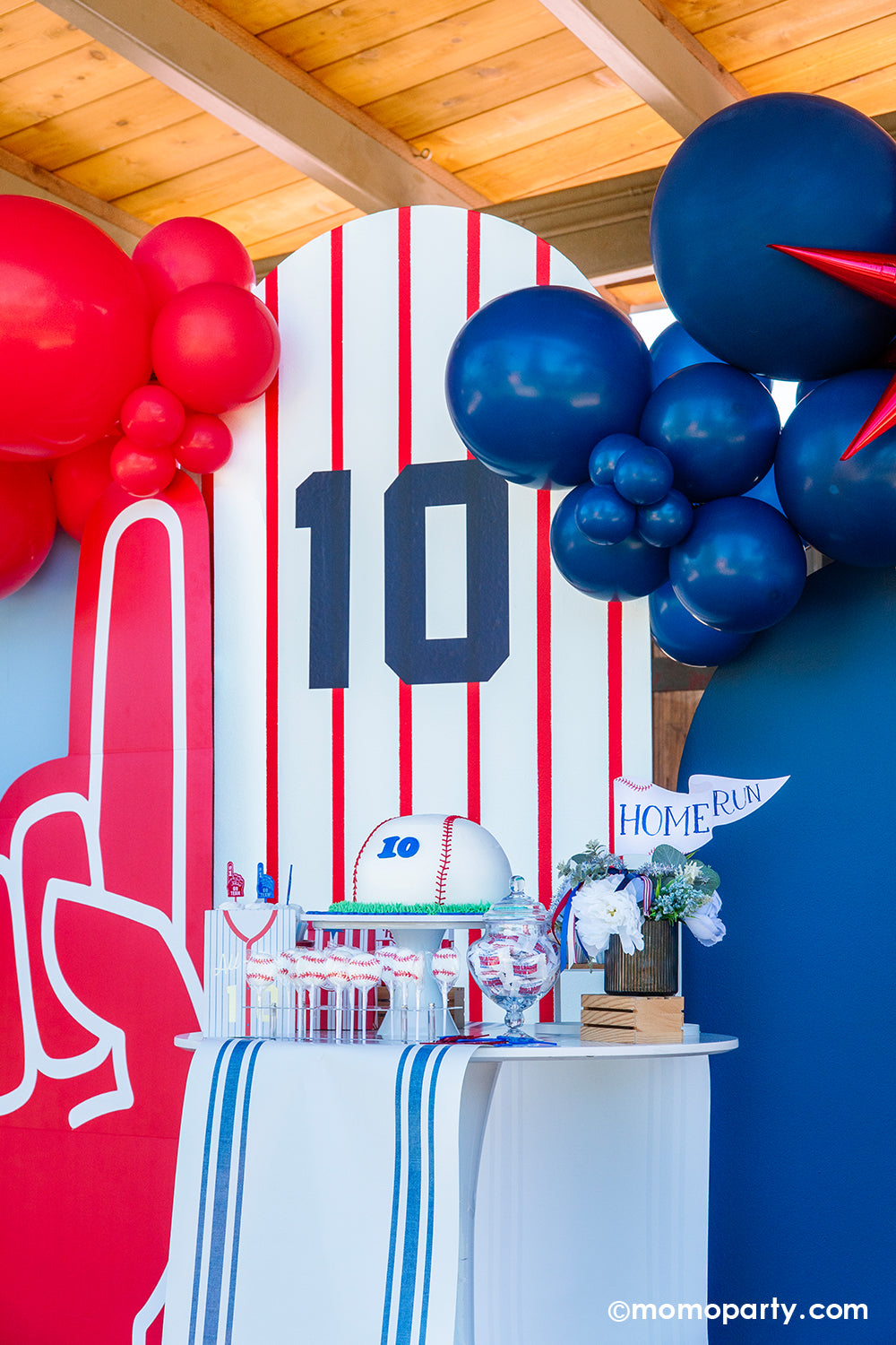 A baseball themed cake and dessert table against a baseball pinstripe backdrop styled by Momo Party featuring a baseball shaped buttercream cake with number 10 on it, cupcakes topped with GO TEAM toppers in blue and red, baseball lollipops, a candy jar with Big League Chews gum balls, and a baseball jersey shaped treat bag, with a floral bouquet and a "Home Run" party pennant as the centerpiece.