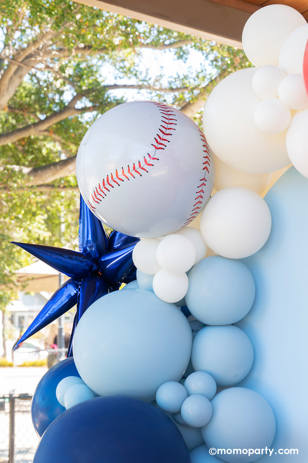 A kid's baseball themed birthday party backdrop decorated with a balloon garland in the classic baseball colors of blue, red and white adorned with a blue starburst foil balloon and a baseball shaped orbz foil balloon from Momo Party.
