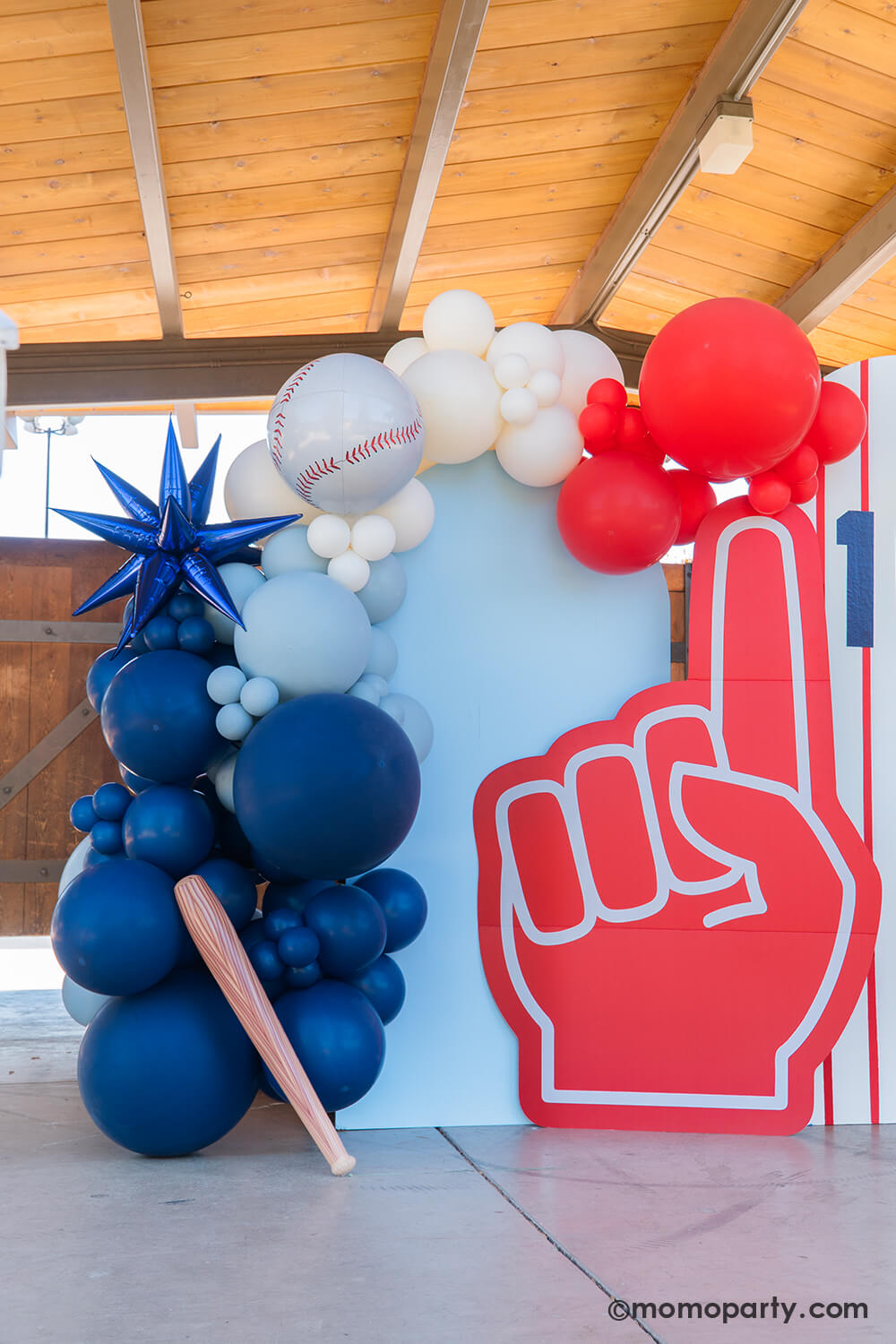 A kid's baseball themed birthday party backdrop decorated with a balloon garland in the classic baseball colors of blue, red and white adorned with a blue starburst foil balloon, a baseball orbz foil balloon a baseball bat shaped inflatable from Momo Party. The party backdrop features red pinstripe design and a foam finger cutout, making this a festive yet modern decoration for a fun celebration for sports.