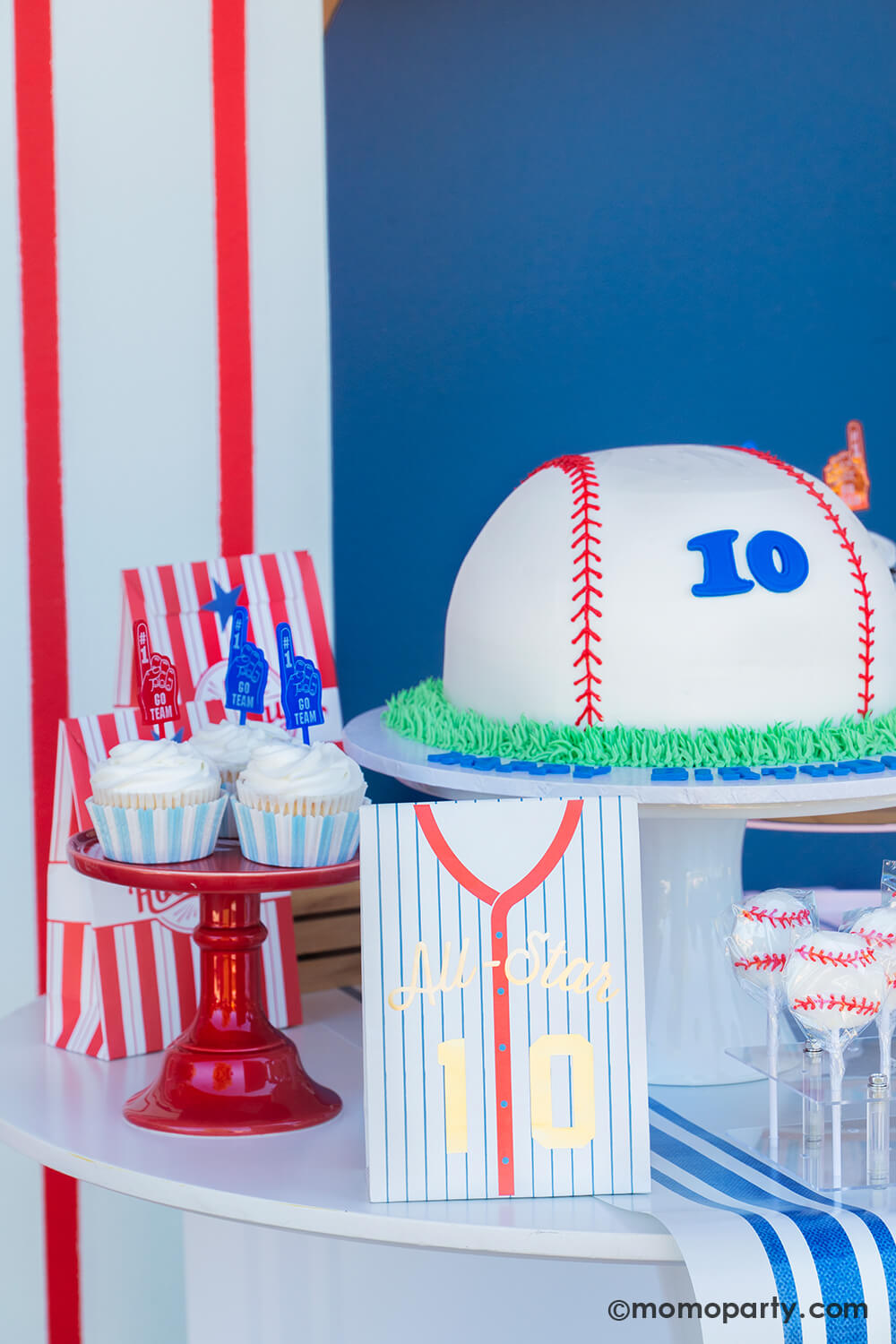 A baseball themed cake and dessert table against a baseball pinstripe backdrop styled by Momo Party featuring a baseball shaped buttercream cake with number 10 on it, the Hip Hip Hooray red striped goodie bags, cupcakes topped with GO TEAM toppers in blue and red, and baseball lollipops.