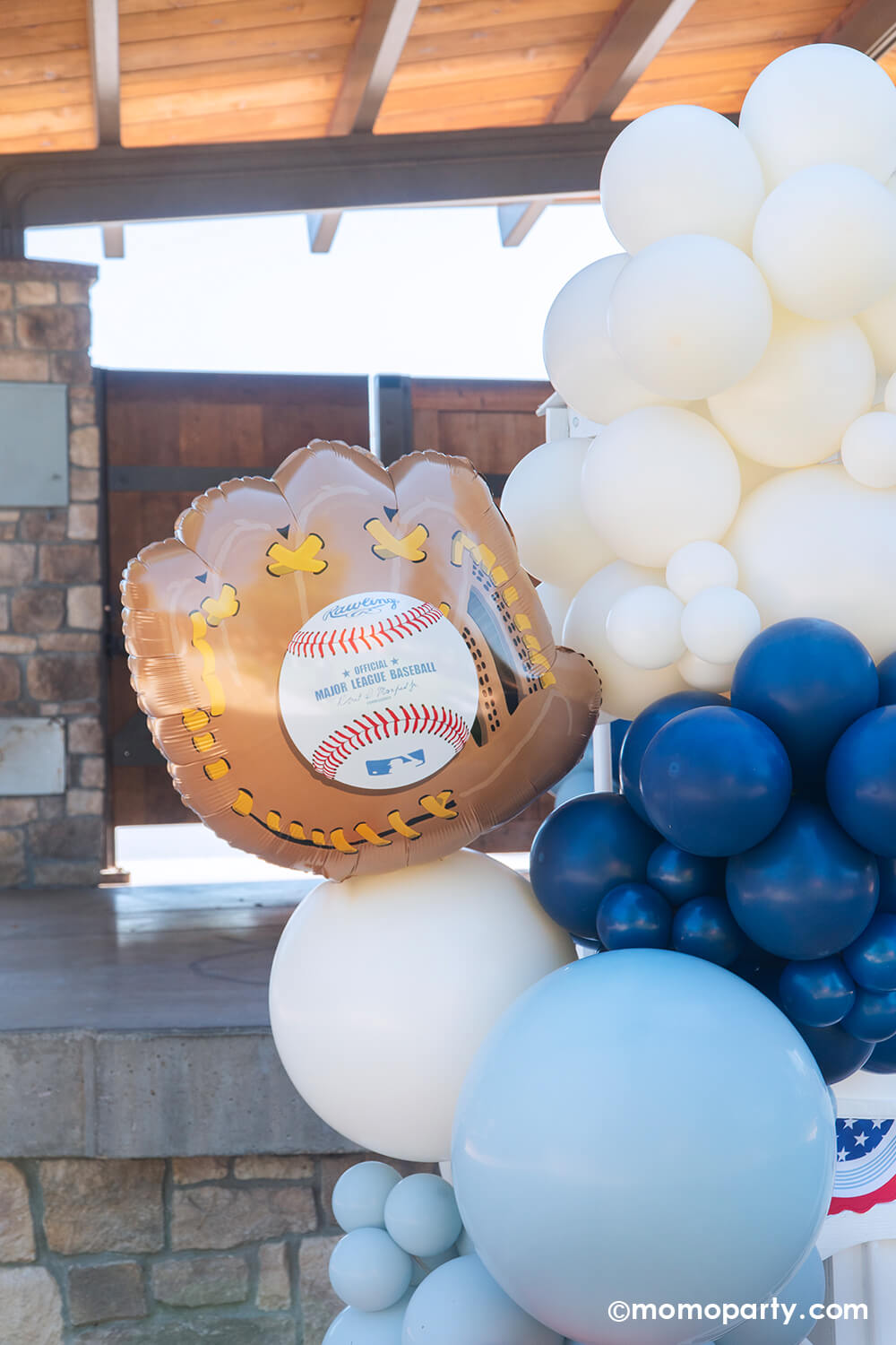 A closeup shot of a candy cart with baseball ballpark snacks decorated with baseball themed balloons in red, blue and white and all-star bunting garland adorned with MLB glove shaped foil balloon from Momo Party at a kid's baseball themed birthday party in a park.