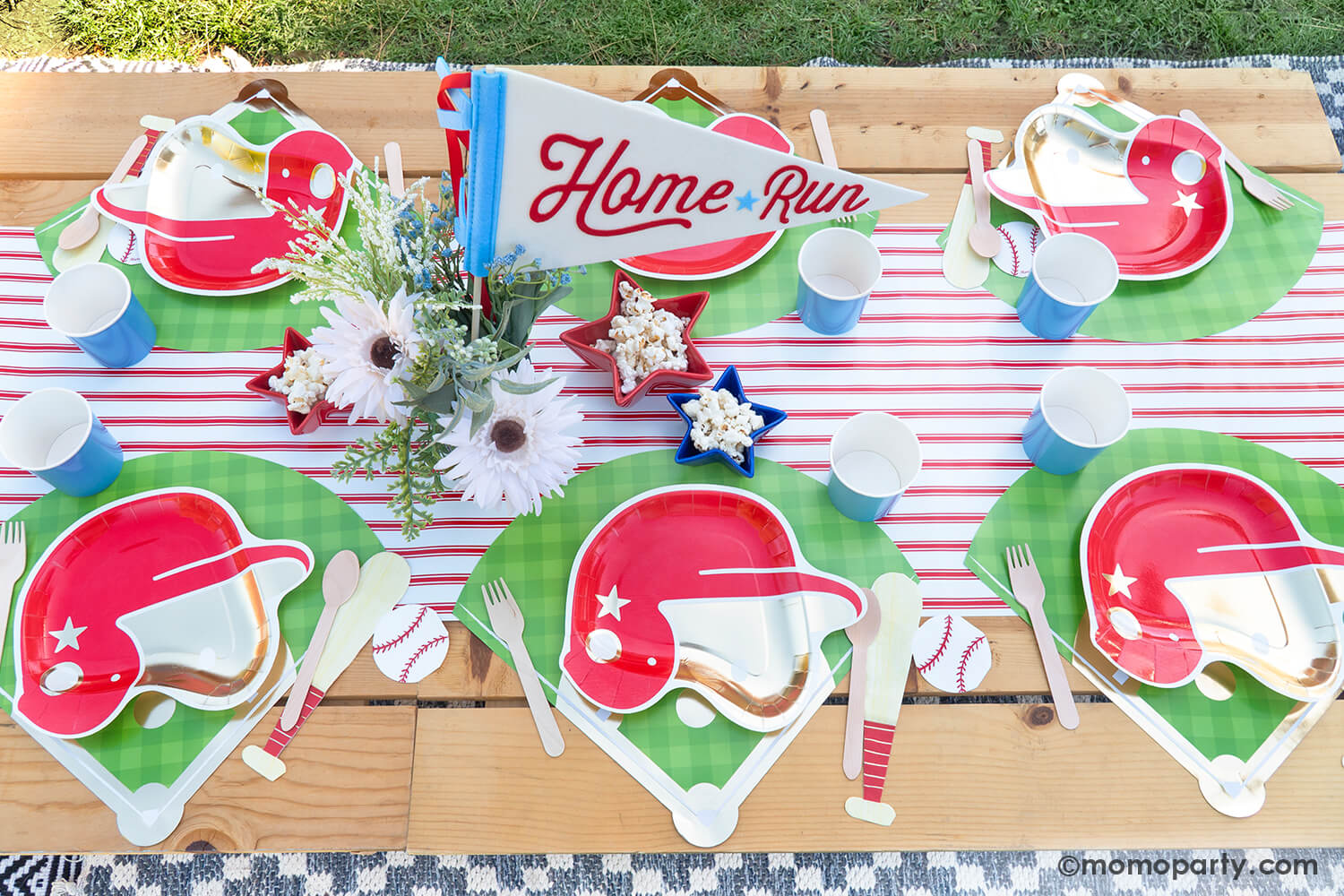 An overhead shot of kid's baseball themed birthday party picnic table setting styled by Momo Party featuring various baseball themed tablewares from Momo Party including baseball helmet shaped plate, cornflower tumbler cups, baseball shaped napkins on the baseball diamond placemats with the red pinstriped runner underneath. In the center there's a beautiful flower bouquet with the "Home Run" felt party pennant and some star shaped trays in blue and red filled with popcorn.