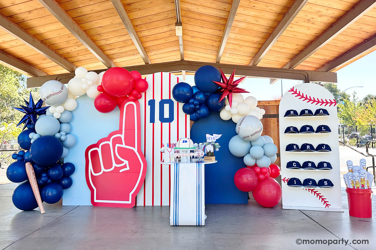 A kid's baseball themed birthday party set up styled by Momo Party featuring a festive yet modern backdrop featuring blue, red and white balloons, A kid's baseball themed birthday party backdrop decorated with a balloon garland in the classic baseball colors of blue, red and white, adorned with a blue starburst foil balloon, a baseball orbz foil balloon a baseball bat shaped inflatable from Momo Party. The party backdrop features red pinstripe design and a foam finger cutout, and a cap wall.