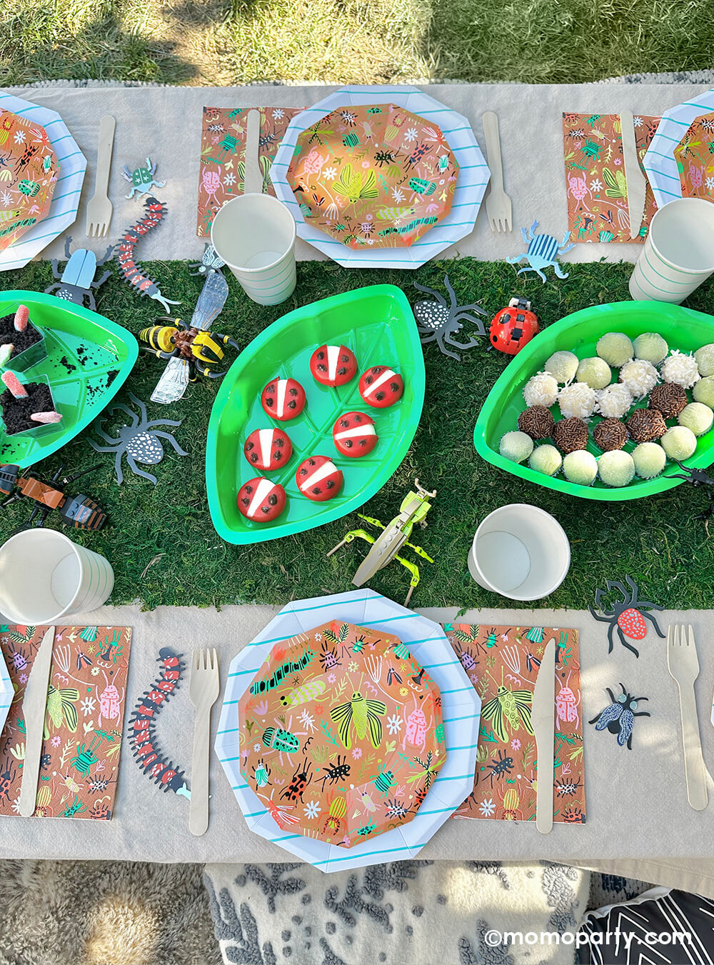 An insect bug themed birthday party table styled by Momo Party features backyard bug plates, napkins with aqua striped plates and party cups by Daydream Society. In the middle there's a faux grass table runner that was decorated with various lego insect toys and palm leaf shaped serving tray filled with insect themed treats and snacks including gummy worms, ladybug babybel cheeses and bug egg shaped brigadeiros, some playful and fun ideas for kid's insect themed bash.