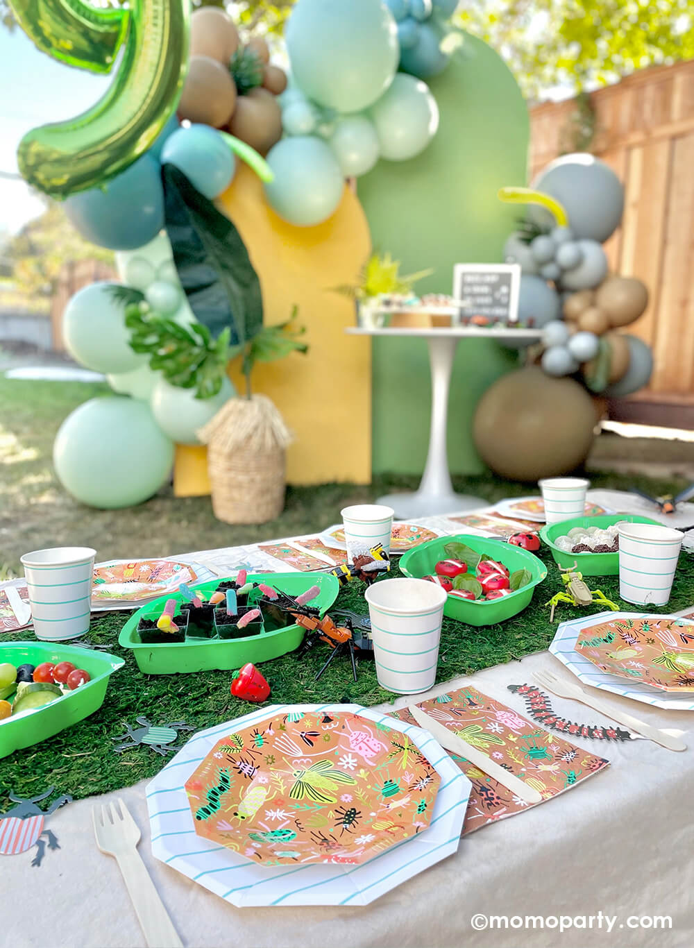 An insect bug themed birthday party table styled by Momo Party features backyard bug plates, napkins with aqua striped plates and party cups by Daydream Society. In the middle there's a faux grass table runner that was decorated with various lego insect toys and palm leaf shaped serving tray filled with insect themed treats and snacks including gummy worms, ladybug cheeses, with a balloon backdrop in green, yellow and brown setting a natural tone for this bug themed party.