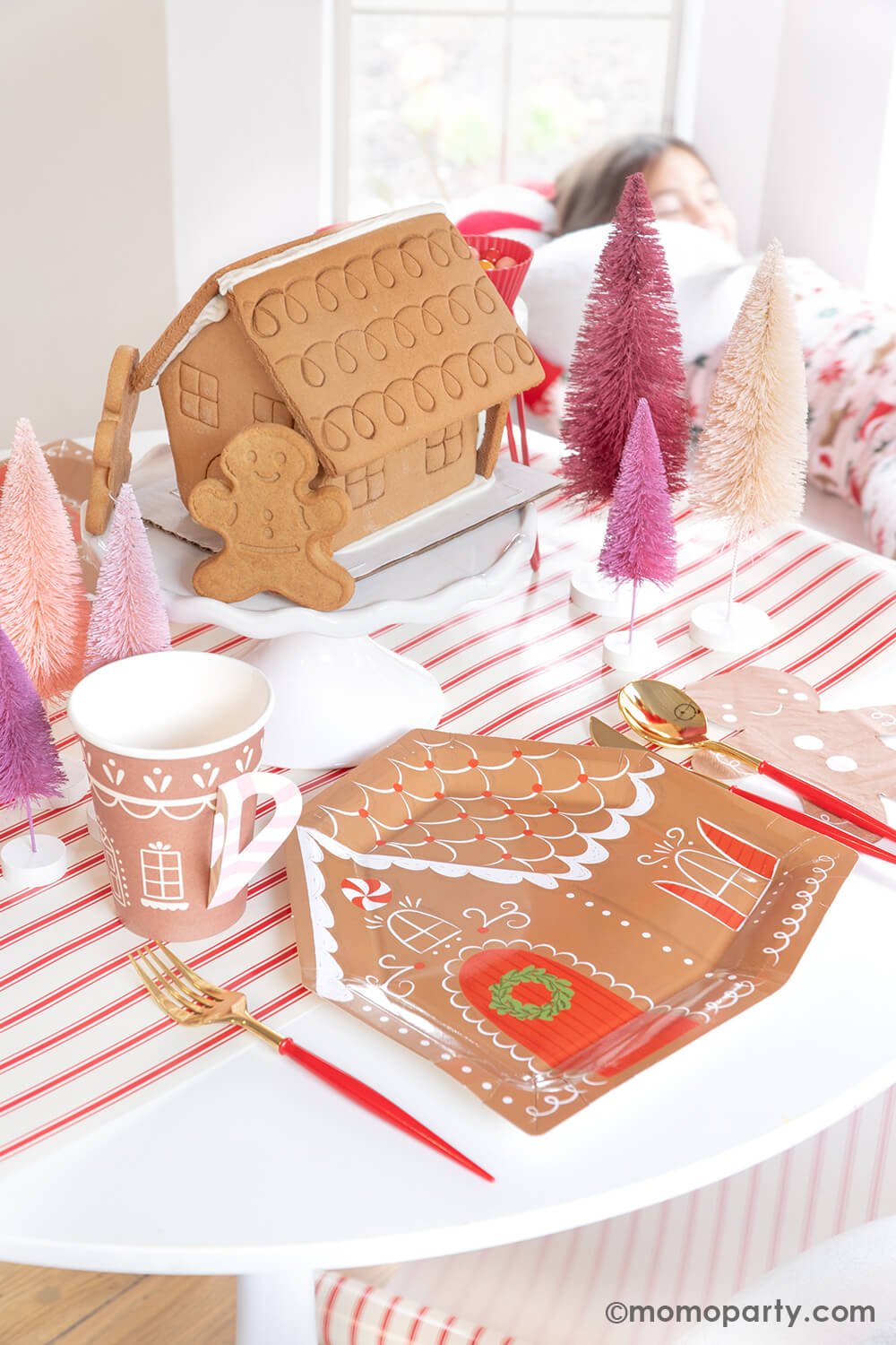 A delightful scene at Momo Party's Gingerbread House Decoration Home Party: A dining table adorned with My Mind's Eye Gingerbread House-Shaped Plates, Smiley Gingerbread Man Napkins, Gold and Red Cutlery Set, and Gingerbread House Paper Party Cups with Handles. Also, Gingerbread House cookies are showcased on a cake stand, surrounded by Christmas Bottle Brush Trees, all set atop the festive Believe Christmas Red Striped Table Runner. What a fun idea to celebrate your holiday with kids at home.