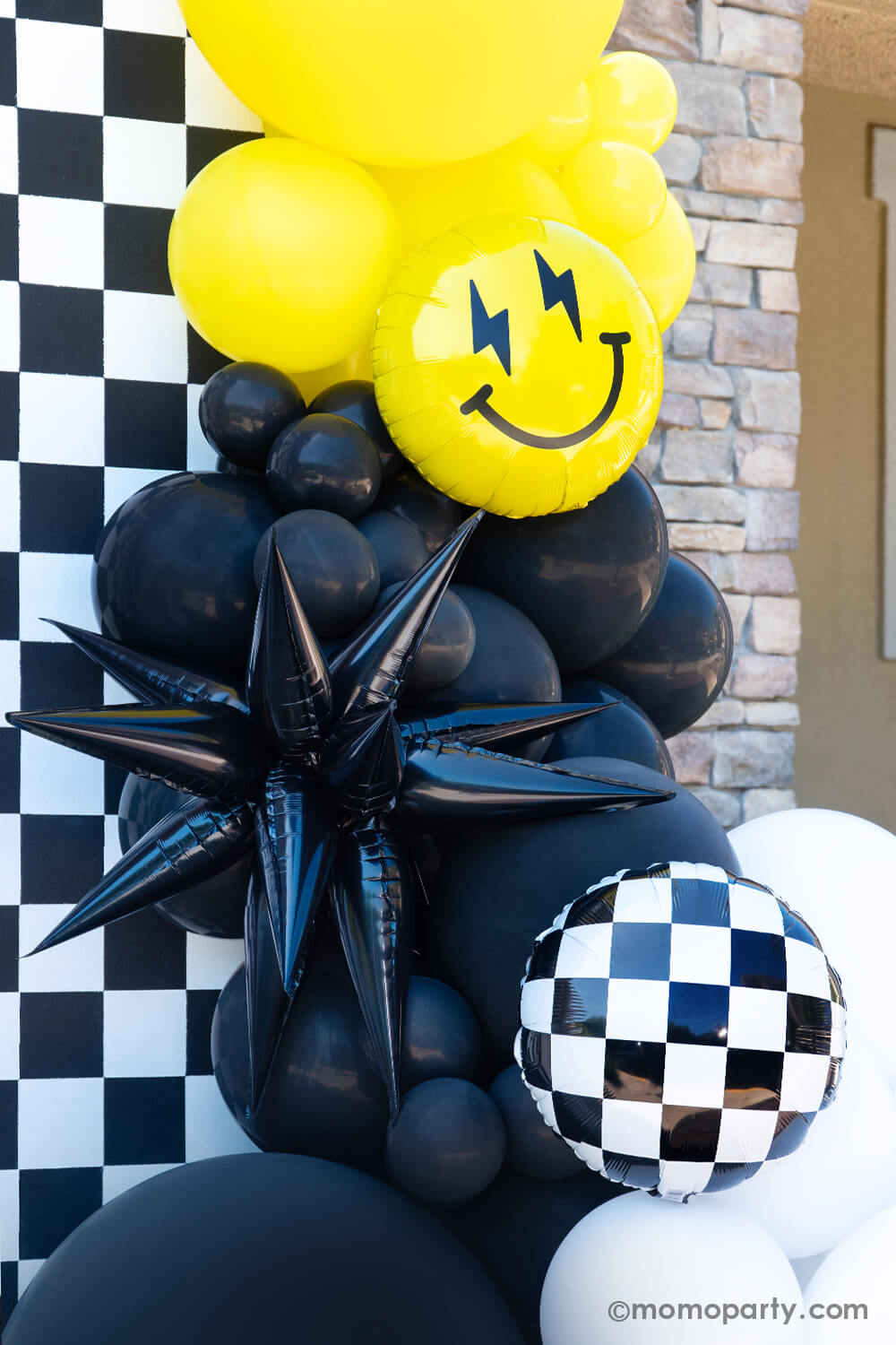 A close up look of Momo Party's "Cool Dude" themed Birthday Party, featuring a balloon garlands in black, white and yellow adorned with a black starburst foil balloon, Checkerboard Foil Balloon and smiley fact foil balloon with lightning bolt eyes, in front of the black and white checkerboard. This decoration is perfect for boy's "One Happy Dude" themed first birthday, a "Two Cool" themed second birthday party or "Ten Rad Years" 10th birthday party.