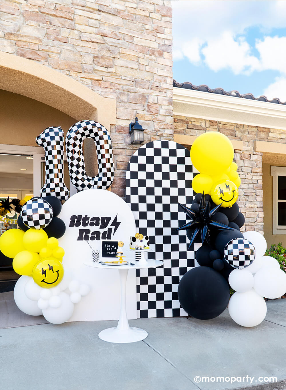 Momo Party's 'Cool Dude' 10th Birthday bash features a stylish balloon garland in black, white, and yellow, starburst and smiley face foil balloons, and Large Number 10 Checkered Foil Balloons. Against a backdrop of checkerboard and a 'Stay Rad' message, the table dazzles with Smiley Gold Foiled Plates, Check It! Checkered Dinner Plates, Checkered Flag Party Cups, and Black Checkerboard Straws. Complete with a dotted fondant cake, it's a cool 'Ten Rad Years' celebration!
