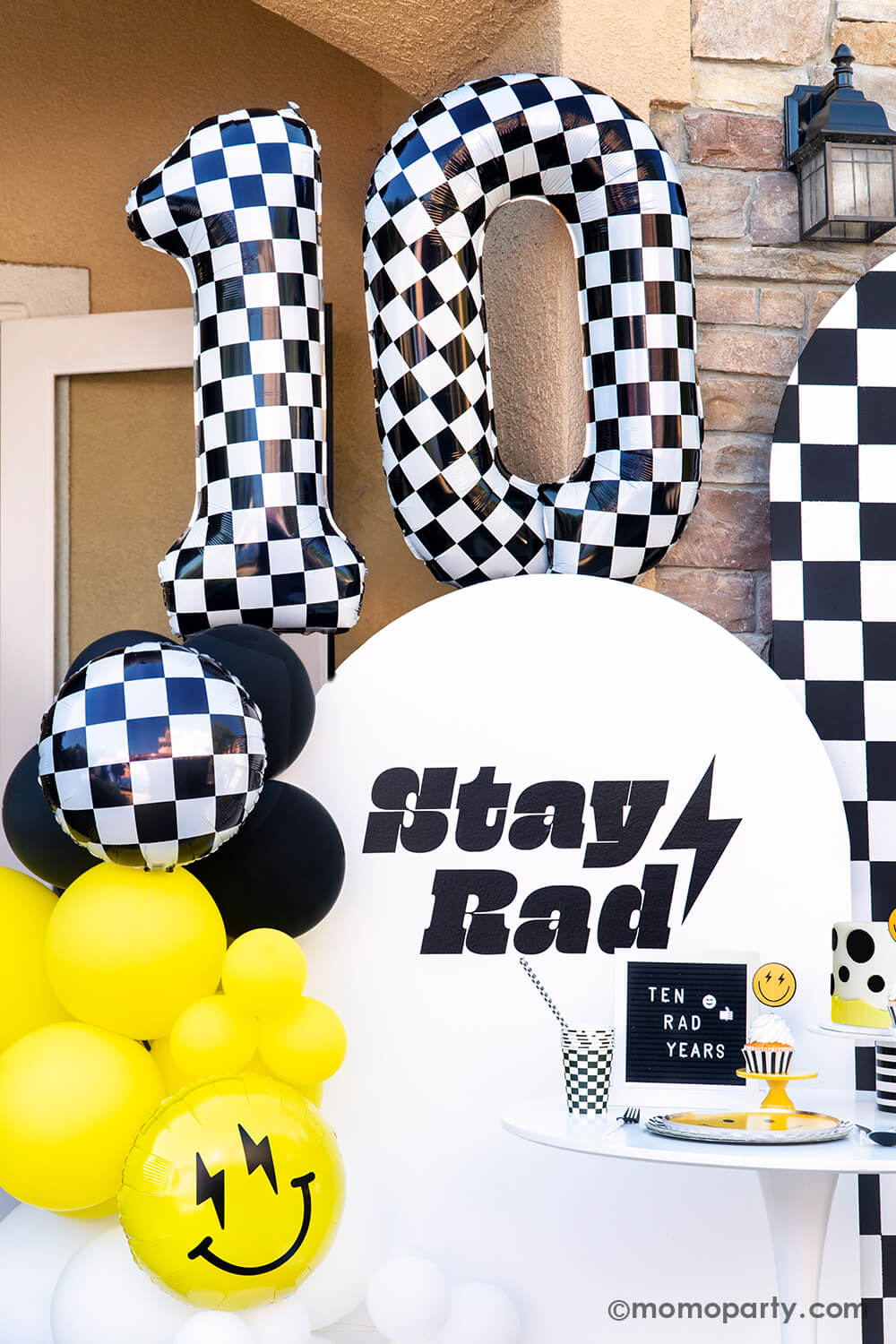 Close up of Momo Party's 'Cool Dude' 10th Birthday bash features a stylish balloon garland in black, white, and yellow, starburst and smiley face foil balloons, and Large Number 10 Checkered Foil Balloons. Against a backdrop of checkerboard and a 'Stay Rad' message, the table dazzles with Smiley Gold Foiled Plates, Check It! Checkered Dinner Plates, Checkered Flag Party Cups, and Black Checkerboard Straws. Complete with a dotted fondant cake, it's a cool 'Ten Rad Years' celebration!
