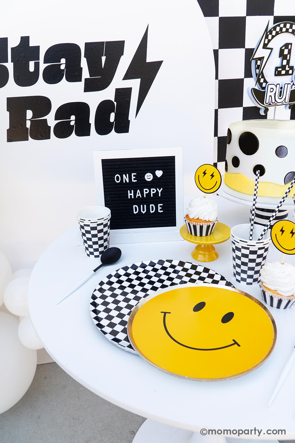 Baby boy's "One Happy Dude" first birthday party tablescape by Momo Party. Featuring the classic black and white checkered patterned plates, cups, napkins and smiley face plates, this rad party table decoration is perfect for your little cool dude's very first big day! With a mix of cool dude aesthetics, smiley-face accents, and checkered patterns in black and white, we've got the perfect recipe for a memorable boy's birthday bash. Don't miss out on making this party as special as your little boy.