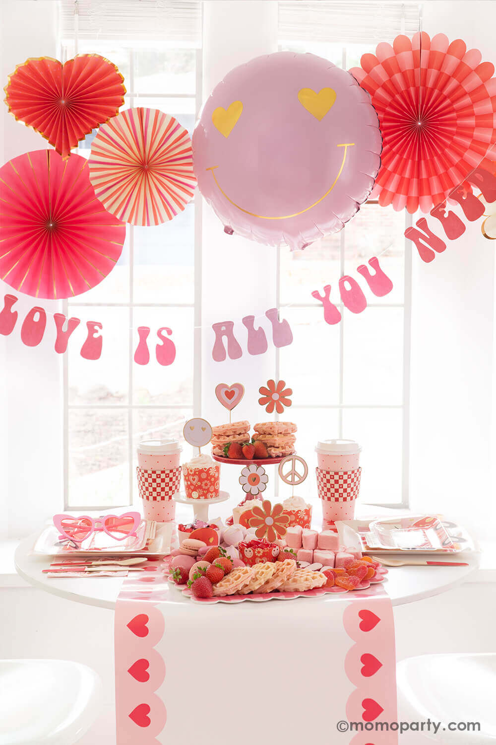 A Valentine's Day party featuring Momo Party's Groovy Valentine's Day Party box set up with pink and red paper fans in heart shape and a "love is all you need" banner in retro type hung below the paper fans. On the table you can see a sweet board featuring treats and snacks in red and pink colors including heart shaped waffles, strawberries, macarons, marshmallows etc. Next to the desert board featured retro inspired Valentine's plates, party cups and napkins, a perfect inspo for Vday celebration!