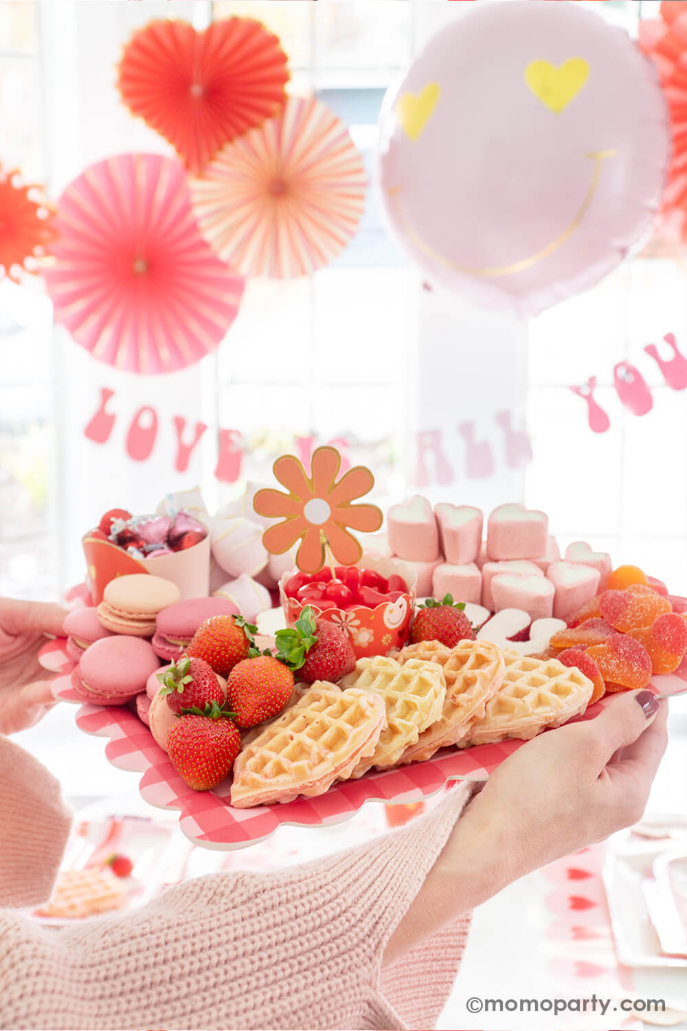 Momo Party presents a scene of Valentine's celebration, featuring a delightful setup where the party host lady holds a My Mind's Eye Checkered Heart-Shaped Tray. The tray is filled with a tempting array of Valentine's Day treats, including "I Love You" sugar cookies, macarons, strawberries, heart-shaped marshmallows, heart-shaped pink waffles, and candies. This charming display serves as the perfect inspiration for a Valentine's Day Dessert Charcuterie Board