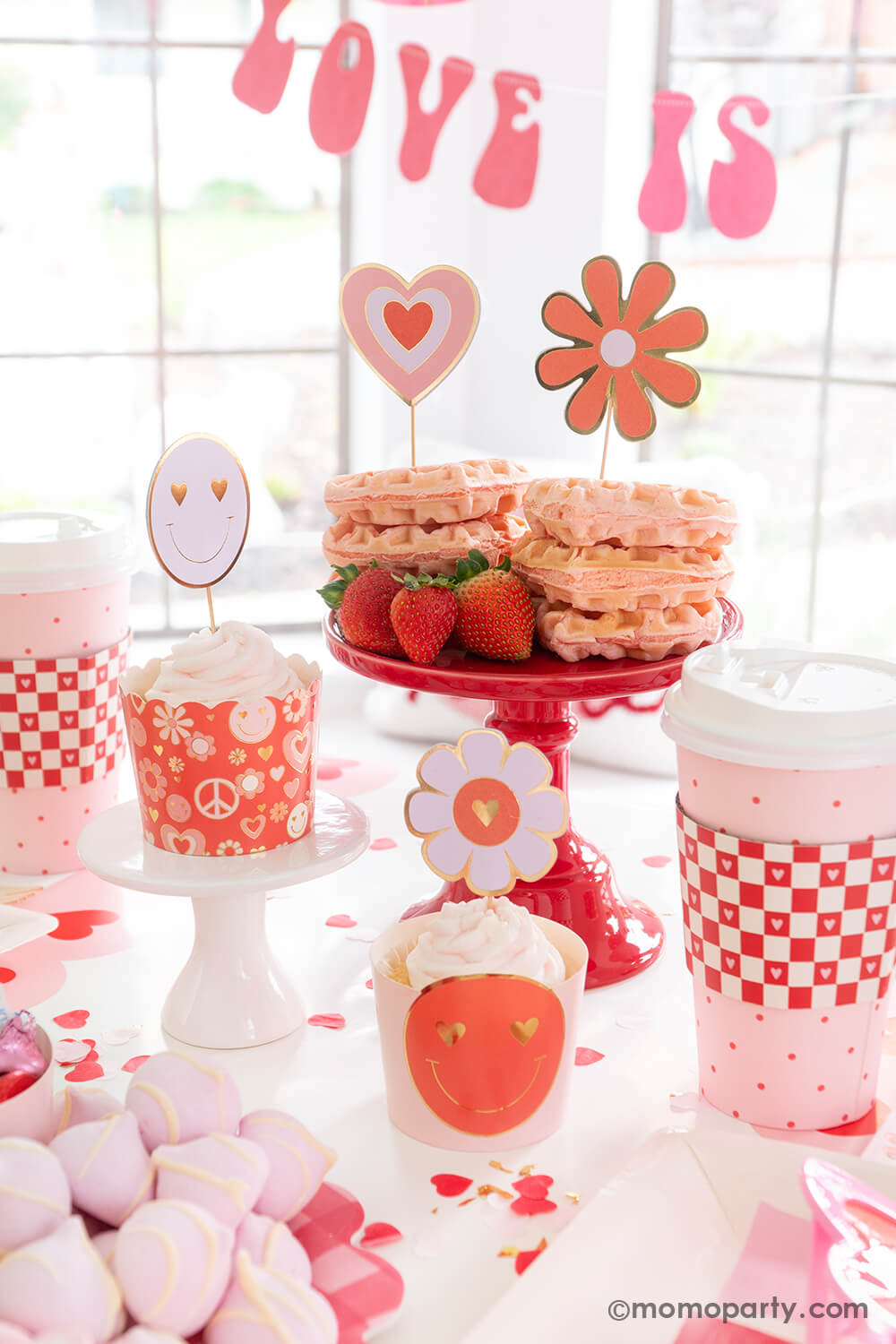 A close up shot of Momo Party's Groovy Valentine's party box featuring My Mind's Eye's love baking cups and toppers set in retro design of daisies, peace signs, smiley faces and hearts. In the back are some heart shaped pink waffles topped with retro toppers with strawberries on the side. Above the table hung "love is all you need" party banner in retro font type, with retro inspired tableware, it makes a perfect inspo for a fun and groovy Valentine's Day celebration!