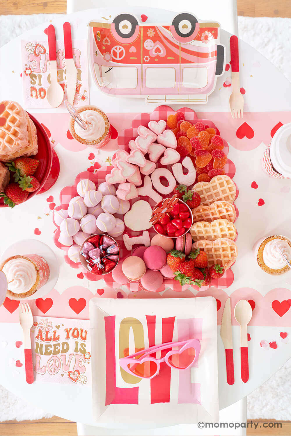 A party table featuring Momo Party's Groovy Valentine's party box with My Mind's Eye's retro Valentine's party supplies including LOVE square plates, VW bus shaped plates, baking cup and topper set and "all you need is love" napkins. On the table there's a heart shaped sweet board filled with Valentine's Day themed treats including "I Love You" sugar cookies, macarons, strawberries, heart shaped marshmallows, heart shaped pink waffles and candies, a perfect inspo for a fun Valentine's celebration!