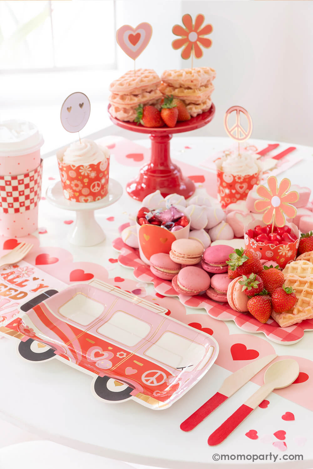 A party table featuring Momo Party's Groovy Valentine's party box with My Mind's Eye's retro Valentine's party supplies including a VW bus shaped plates, red checkered party cups, baking cup and topper set and "all you need is love" napkins. On the table there's a heart shaped sweet board filled with Valentine's Day themed treats including macarons, strawberries, heart shaped marshmallows, heart shaped pink waffles and candies, a perfect inspo for a fun Valentine's celebration!