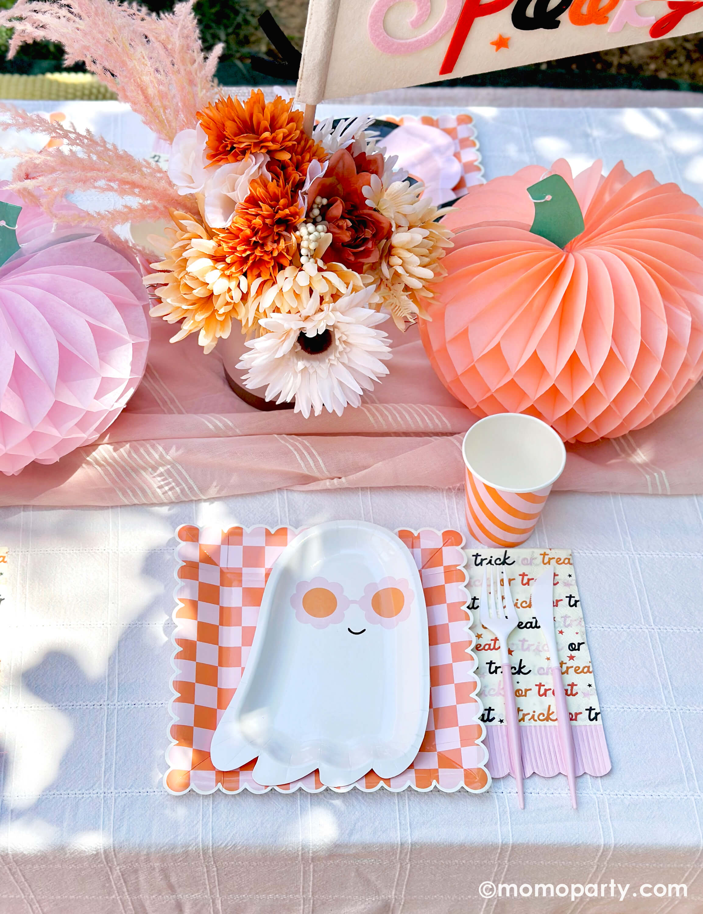 A pink Halloween table featuring Momo Party's pink sunny ghost shaped plates paired with orange/pink checkered plates, with boho vibe fall flower arrangement and pastel peach and pink honeycomb pumpkins as the centerpiece, and a felt party pennant that has "spooky" written on, this tablescape gives a great inspiration for a modern and chic Halloween party table decoration ideas.