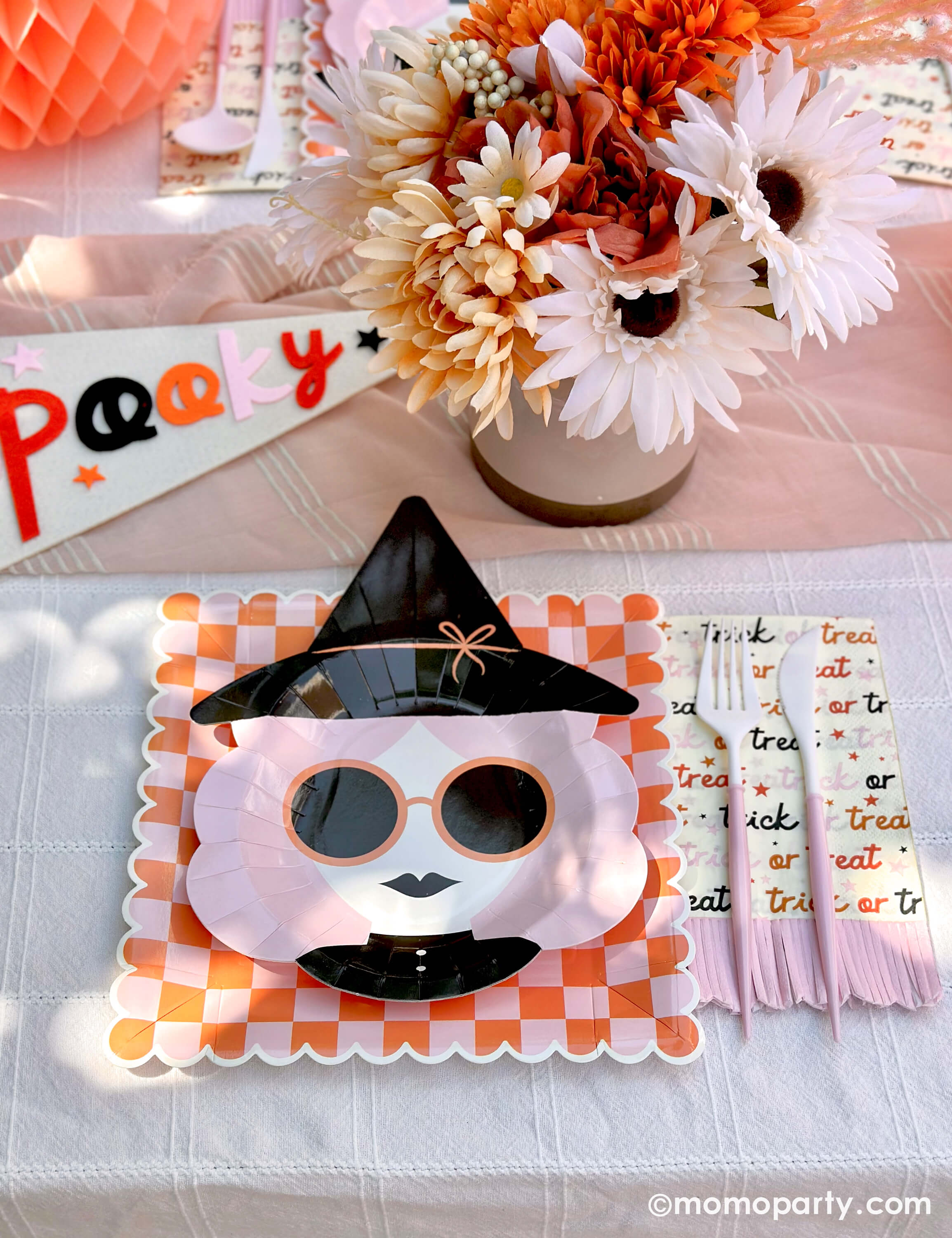A pink Halloween table featuring Momo Party's pink witch plates paired with orange/pink checkered plates, trick or treat patterned guest towels with pastel pink fringe, and blush and white cutlery, with boho vibe fall flower arrangement and pastel peach honeycomb pumpkins as the centerpiece, and a felt party pennant that has "spooky" written on, this tablescape gives a great inspiration for a modern and chic Halloween party table decoration ideas.