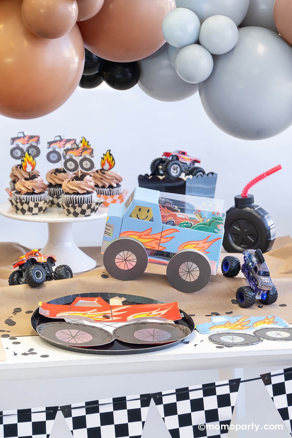 A monster truck themed party table setting styled by Momo Party decorated with party balloon cloud and checkerboard race car flags banner. The table features a red monster truck shaped paper plate with black round dinner plate, a truck shaped blue napkin, the monster truck cupcake kit and brown cupcakes topped with flame toppers, a tire shaped tumbler cup with red straw on a brown craft table cover decorated with monster truck toys and black confetti - a fun setup for kid's birthday party.