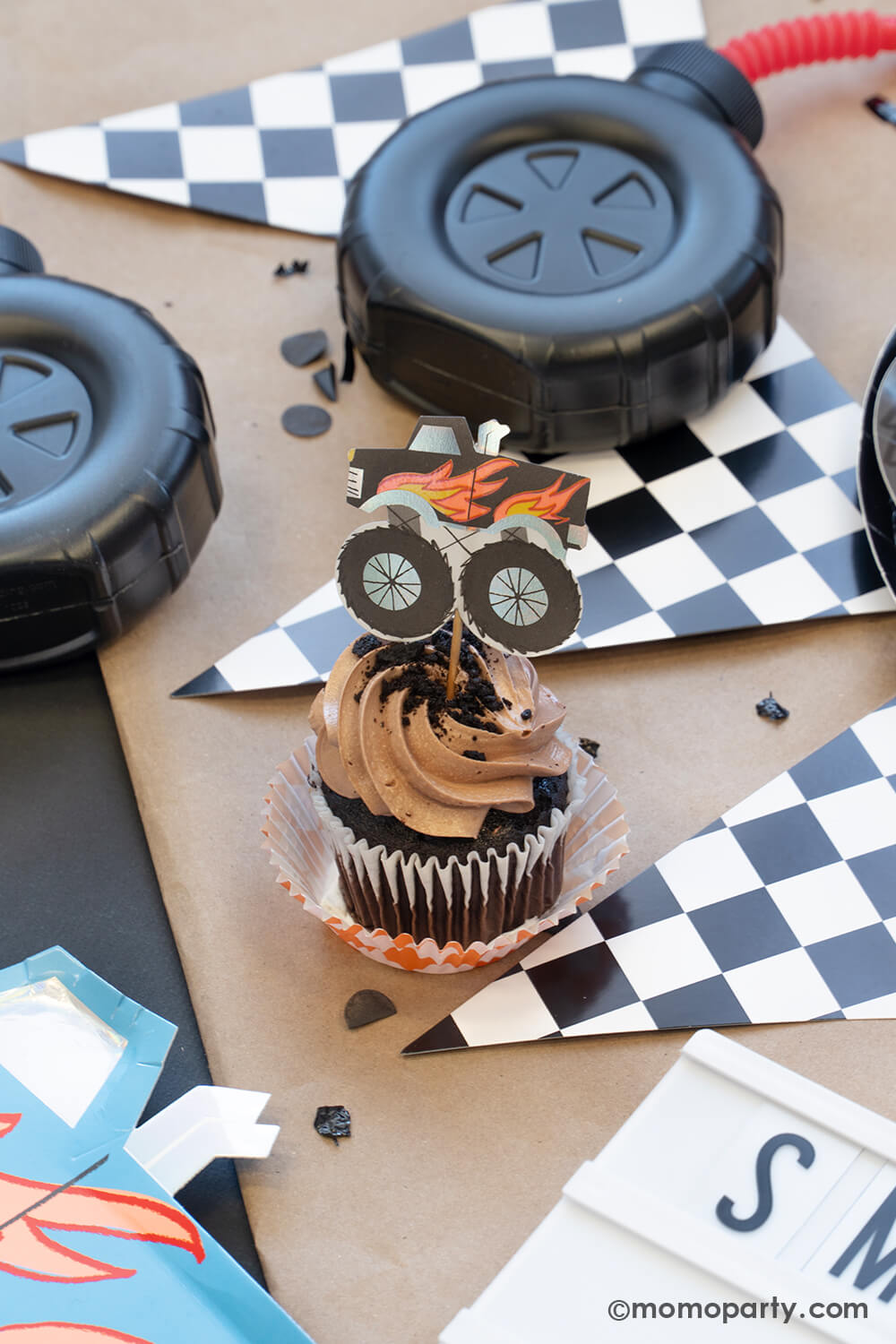 A cupcake with brown icing and oreo crumbs topped with a black monster truck topper from Momo Party. Around the cupcake there's a race car flag checkerboard party banner and a few tire shaped tumblers with red straws, all on a brown kraft paper table runner with black confetti around it. Making this a fun and festive scene for kid's monster truck themed party celebration.
