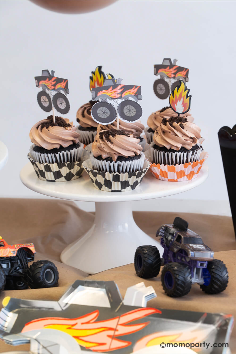 A monster truck themed party table setting styled by Momo Party. The table features a black monster truck shaped paper plate and a few cupcakes with brown icing and oreo crumbs on them, each of them is topped with either a fire flame topper or a black monster truck shaped topper. Next to the cupcakes there's a black popcorn box filled with popcorn with a monster truck toy on it. The table is covered with brown kraft paper with a few colorful monster truck toys on it, fun for kid's birthday celebrations!