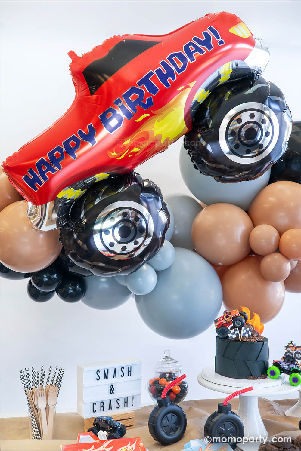 A close up of the monster truck dirt road themed balloon garland featuring various sizes of latex balloons in brown, black and grey colors with the red happy birthday monster truck shaped foil balloon by Momo Party. Underneath the balloon garland is a monster truck themed party table with a large tire shaped birthday cake and a "Smash & Crash" sign, ready for a fun and high-energy filled celebration for the little monster truck lovers!