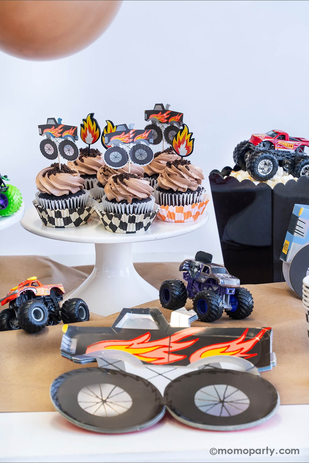 A monster truck themed party table setting styled by Momo Party. The table features a black monster truck shaped paper plate and a few cupcakes with brown icing and oreo crumbs on them, each of them is topped with either a fire flame topper or a black monster truck shaped topper. Next to the cupcakes there's a black popcorn box filled with popcorn with a monster truck toy on it. The table is covered with brown kraft paper with a few colorful monster truck toys on it, fun for kid's birthday celebrations!